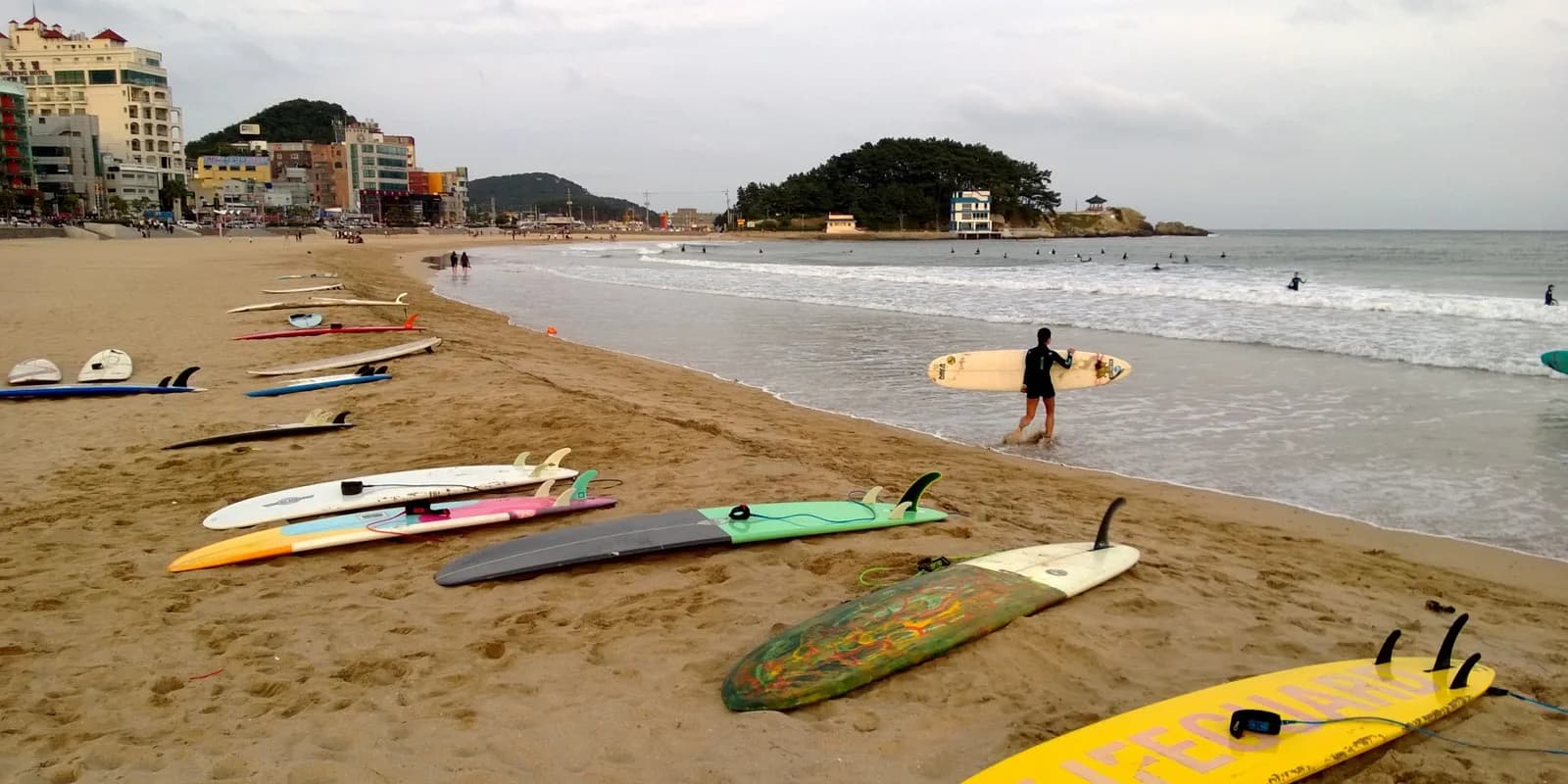 부산 송정해수욕장의 완만한 백사장과 서핑하는 사람들, 오른쪽 끝 죽도공원이 보이는 바다 풍경